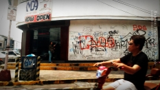 Graffiti. A man on a motorcycle drives by a establishment that has a wall covered by writings.